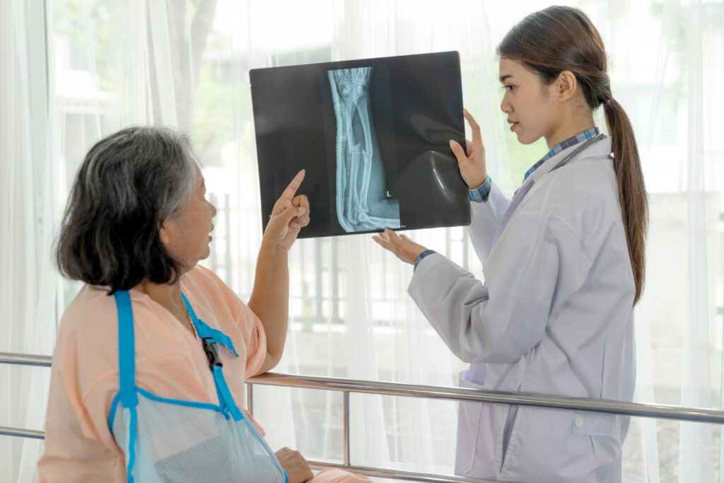 La imagen muestra a dos mujeres en lo que parece ser un hospital. De izquierda a derecha, hay una mujer mayor con cabello grisáceo y ligeramente ondulado. En la foto se puede ver su brazo derecho apoyado en un cabestrillo. A su lado, hay una mujer más joven de cabello castaño, liso y recogido en una cola de caballo. Ella usa una bata blanca y sostiene una radiografía que muestra un hueso roto.