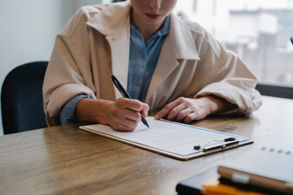 La imagen muestra a una mujer con el rostro parcialmente visible, que viste una camisa formal de color azul debajo de un sobretodo beige y está sentada en una mesa de madera. Con un lapicero negro, toma notas en una hoja sujeta a un portapapeles.