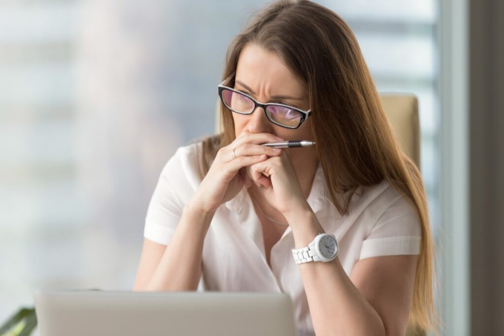 La imagen muestra a una mujer blanca, con el cabello liso y castaño claro, a la altura de los hombros. Lleva una camisa blanca y gafas cuadradas. Está mirando hacia el lado izquierdo de la imagen, sus manos están delante de su boca y está arrugando el entrecejo, como si estuviera pensando. 