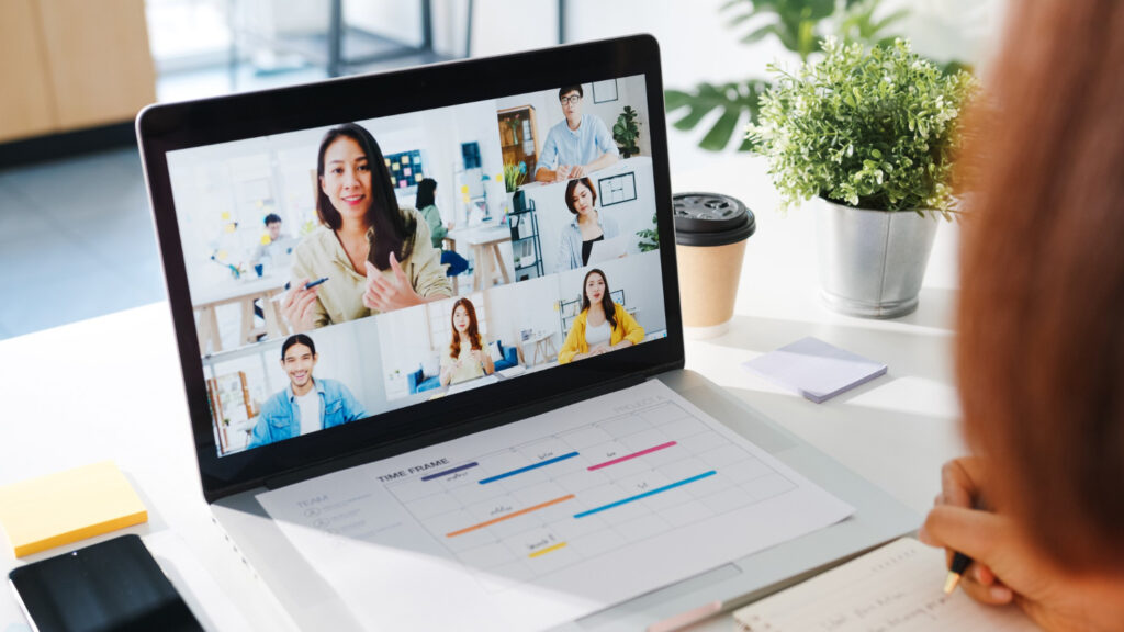 La imagen muestra una laptop plateada sobre la mesa y en la pantalla se puede ver una videollamada con varias personas.