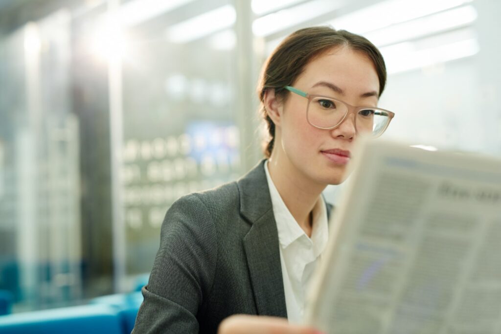 La imagen muestra a una mujer asiática de piel blanca y cabello castaño claro. Usa lentes cuadrados de color marrón claro. La mujer mira un periódico que está delante de ella y aparece desenfocado en la foto. 