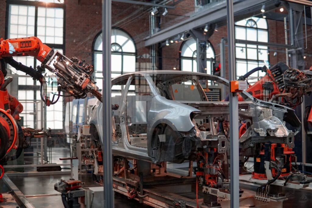 La imagen muestra la carrocería de un auto plateado sostenida por una máquina naranja. En la foto, el auto parece estar en proceso de montaje, todavía en la fábrica. 