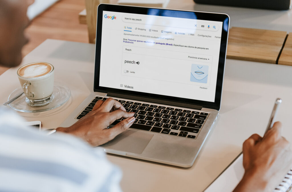 La imagen muestra una laptop plateada sobre una mesa blanca. En la pantalla, se ve el buscador de Google con la palabra “peach” destacada. También se puede ver la mano izquierda de una persona negra en el teclado de la laptop. Su mano derecha está sosteniendo un lapicero sobre un cuaderno de notas.