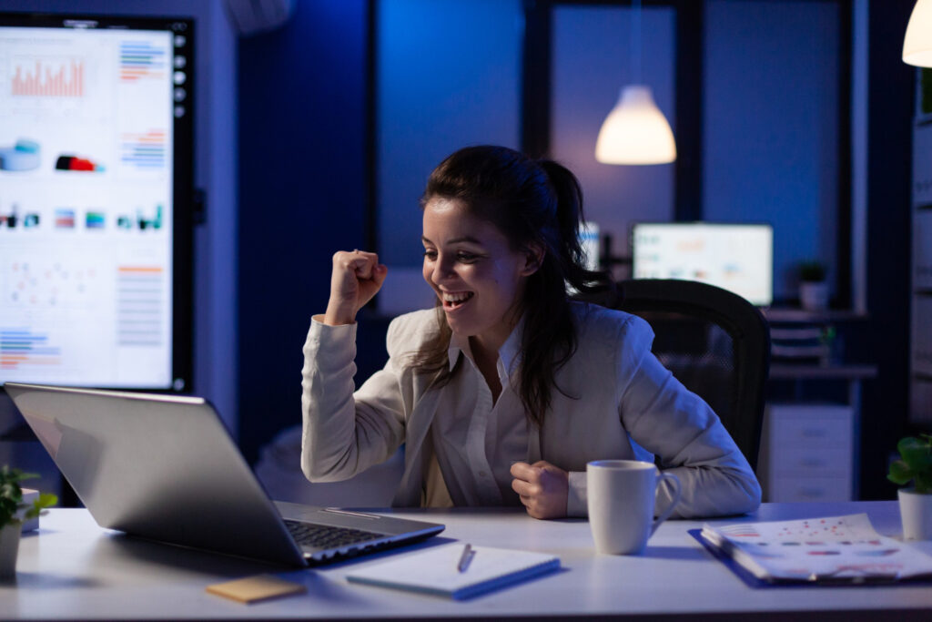Una mujer de cabello castaño está frente a una laptop plateada. Ella está sonriendo y hace un gesto con las manos —una especie de puño en el aire— como si estuviese celebrando algo.