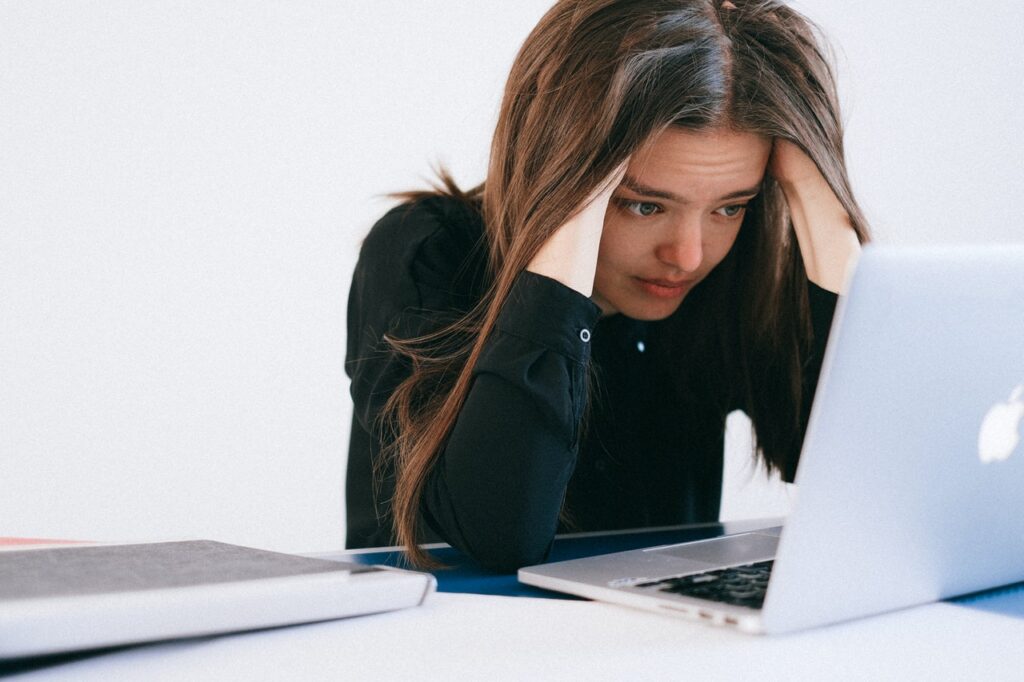 Una mujer blanca de cabello castaño liso está frente a una laptop plateada. Ella mira la pantalla y se agarra la cabeza con sus manos como si estuviese preocupada o insatisfecha.