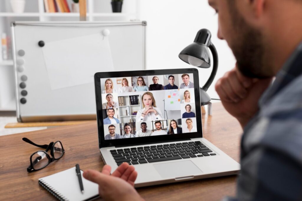 Un hombre blanco está mirando una computadora portátil plateada. En la pantalla se puede ver una videollamada con varias personas.