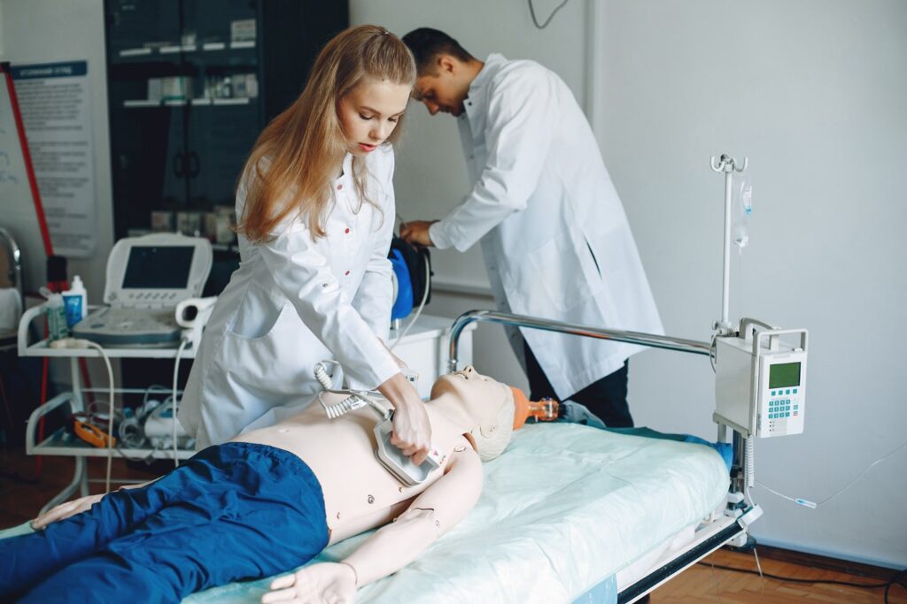Una joven blanca de cabello rubio y liso, vestida con una bata de laboratorio blanca, utiliza un desfibrilador en un maniquí que está en una camilla de hospital. En el fondo, se puede ver a un chico blanco que también lleva una bata de laboratorio blanca.
