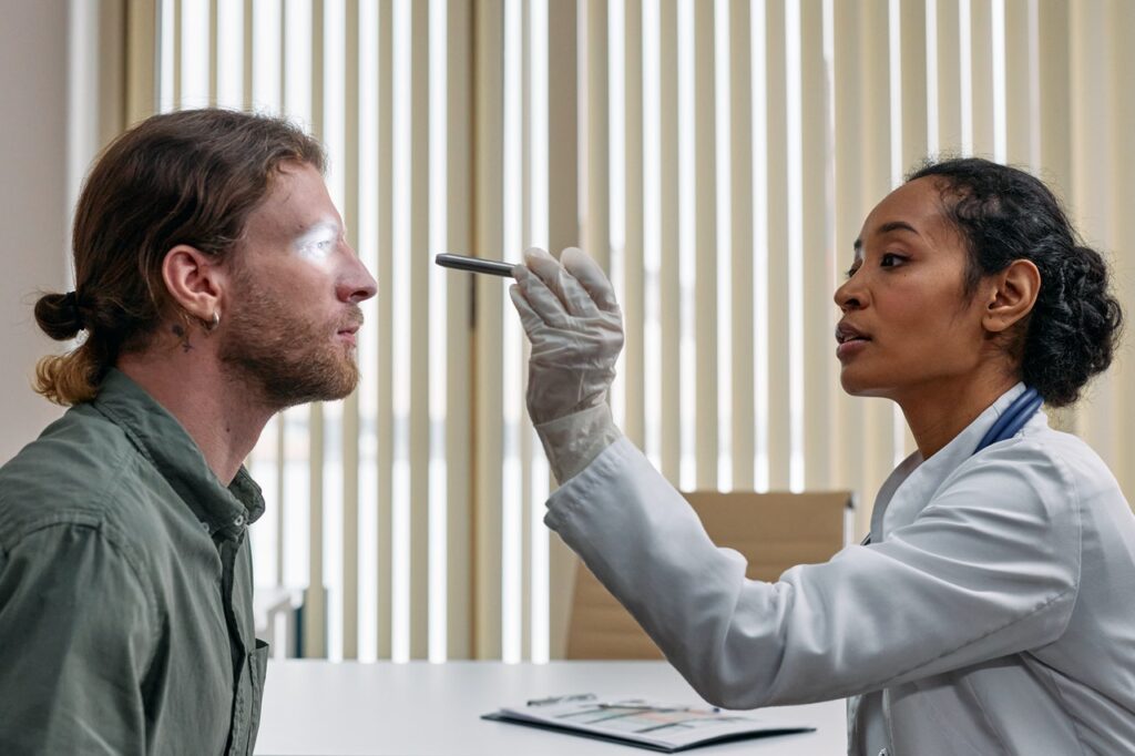 Un hombre blanco con el cabello liso, castaño claro y recogido en un moño se encuentra frente a una doctora de cabello negro. Ella lleva una bata blanca y apunta con una pequeña linterna hacia el ojo del hombre.