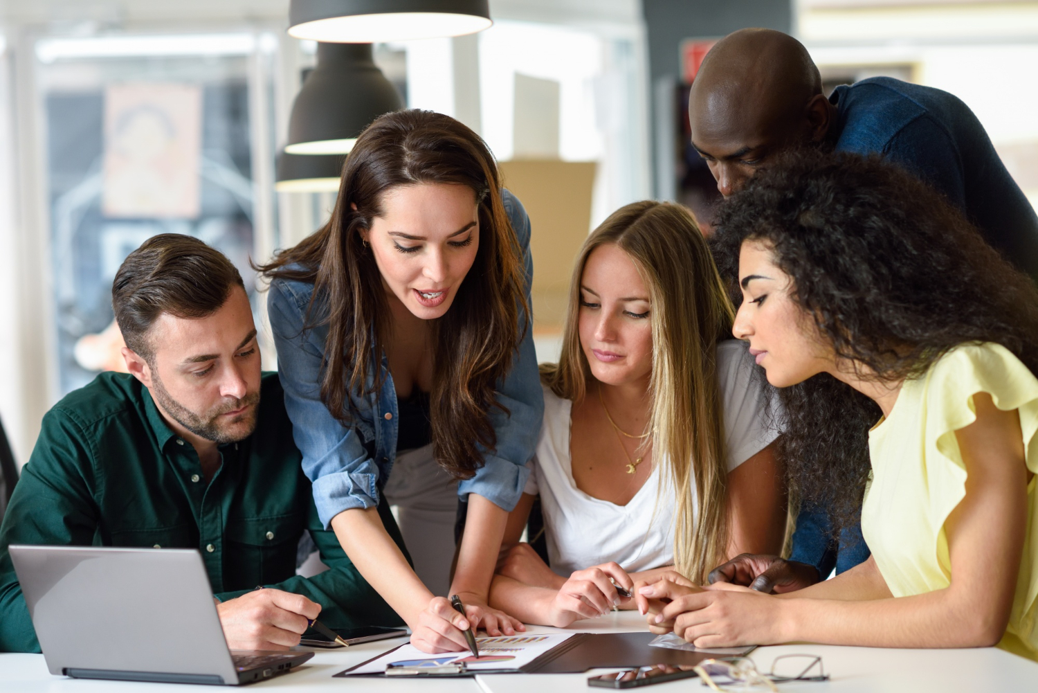 A imagem mostra um grupo de pessoas no ambiente de trabalho em frente a uma mesa. Na foto, é possível ver uma mulher com uma planilha impressa, enquanto as outras pessoas olham para o que ela está mostrando.