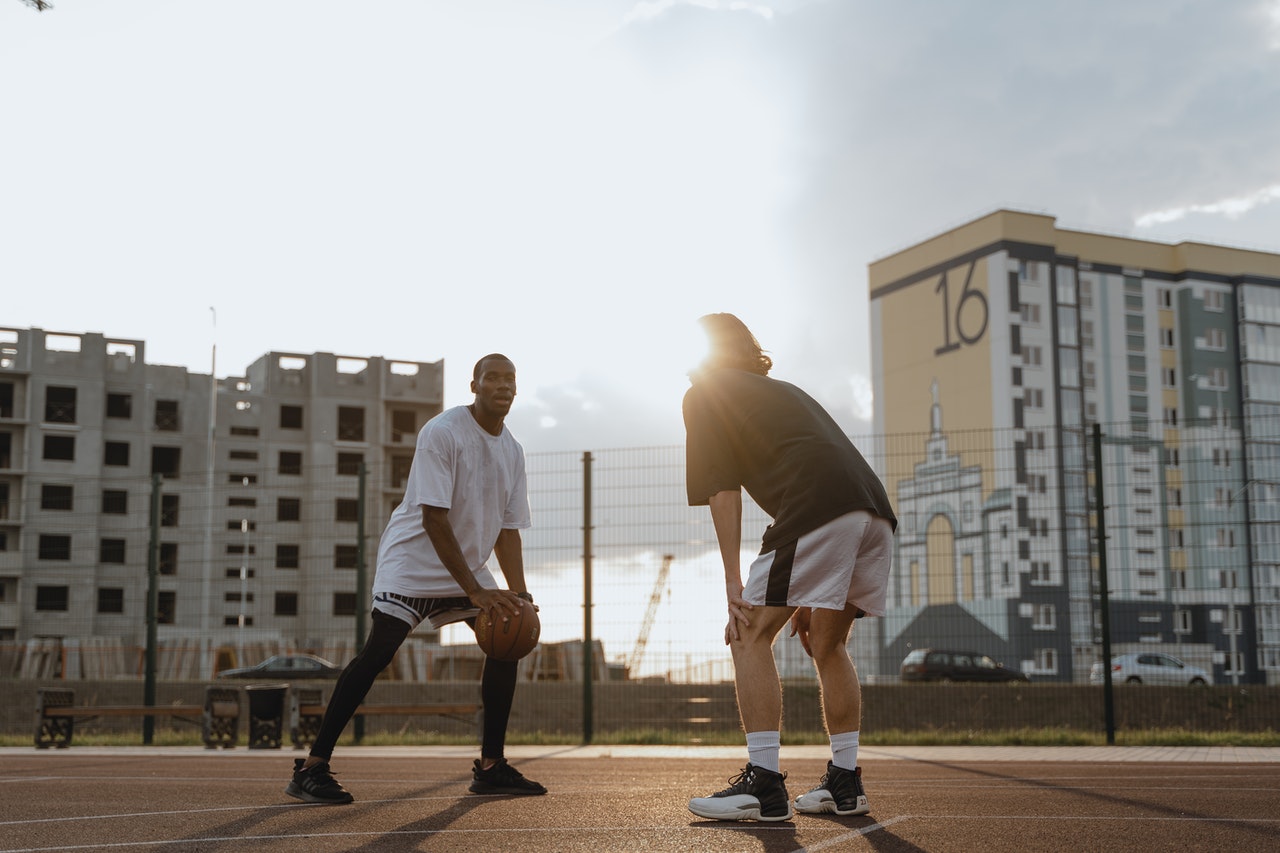 A imagem mostra dois homens em uma quadra de basquete ao ar livre. Da esquerda para a direita, há um homem negro, olhando para a câmera e segurando a bola de basquete. Em frente a ele e de costas para a imagem, está um homem branco, de cabelos até a altura da nuca, que está levemente curvado e com as mãos nos joelhos.