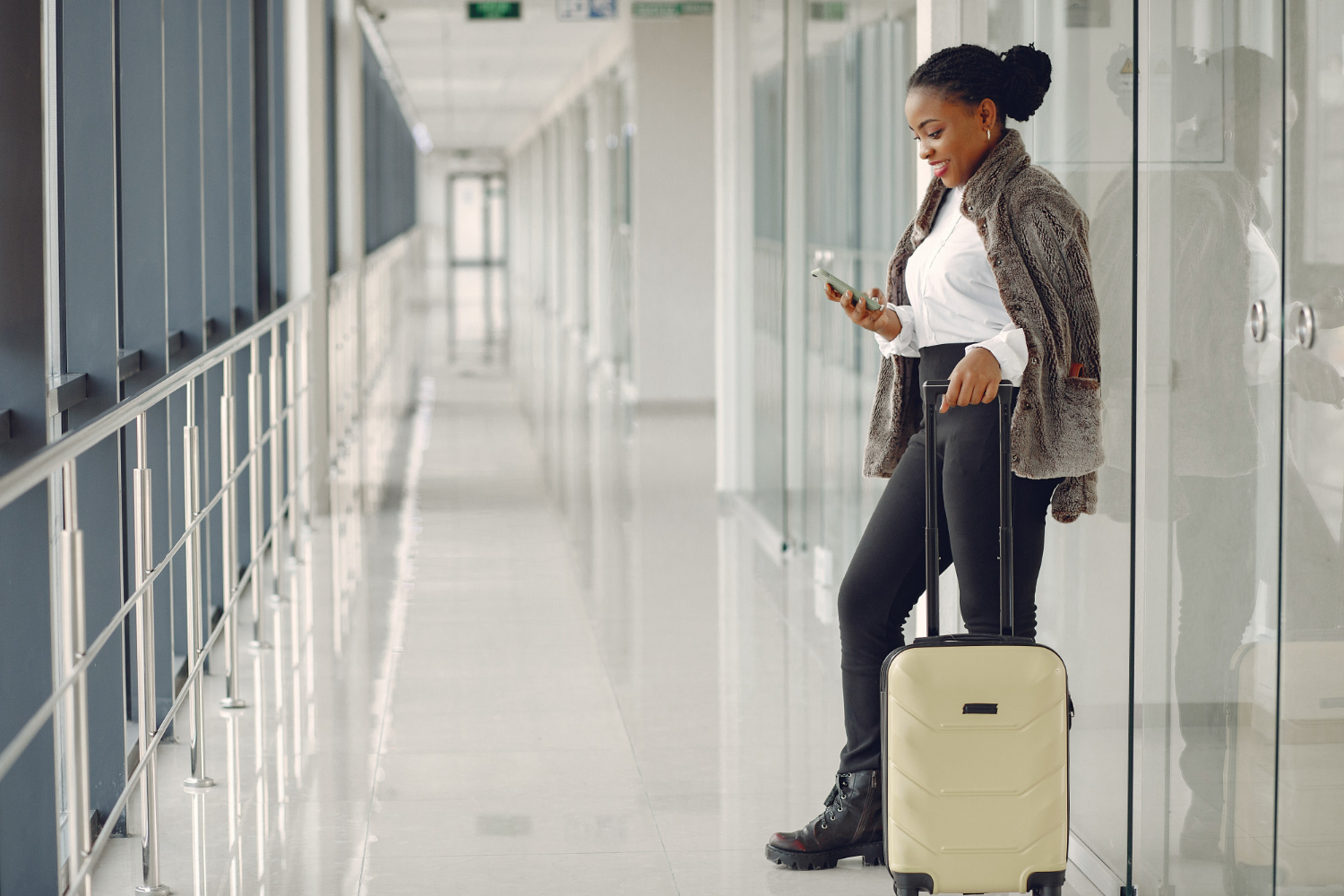 A imagem mostra uma mulher negra que está de lado. Ela está posicionada no canto direito da foto e segura uma mala de viagens de rodinhas na cor creme. 