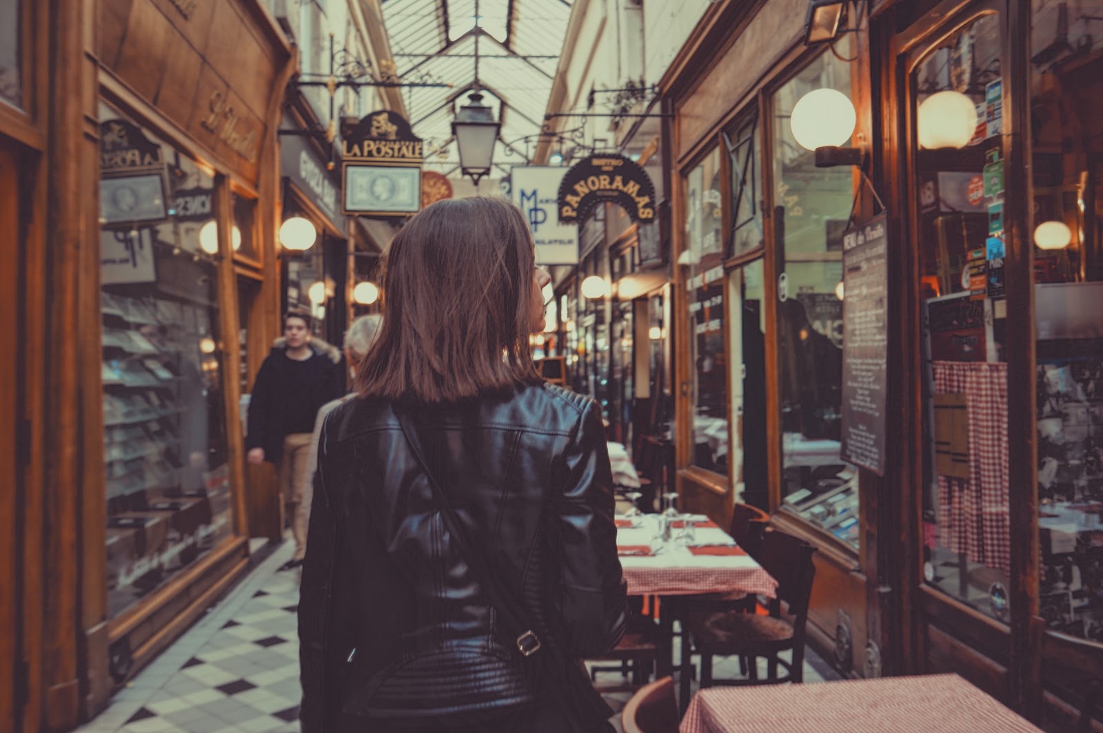 A imagem mostra uma mulher de costas para a imagem. Ela é branca, possui cabelos castanhos lisos na altura do ombro. Essa mulher está caminhando em um corredor de lojas e é possível ver algumas mesas de um restaurante perto dela. 