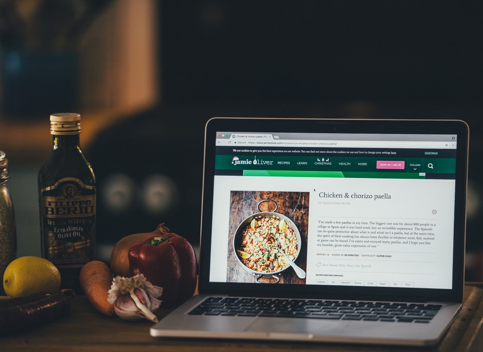 A imagem mostra um notebook prata sob uma mesa de madeira, que está na cozinha. Na tela dele, é possível ver uma receita em inglês. Ao lado esquerdo do notebook, estão alguns ingredientes, como alho, pimentão, cenoura, cebola e limão.