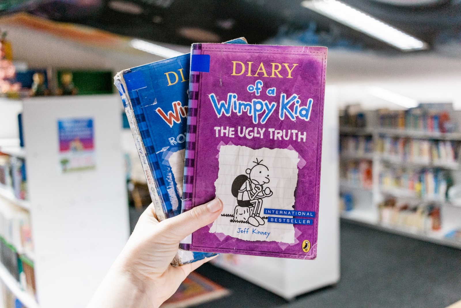 A imagem mostra a mão esquerda de uma pessoa segurando dois livros. O livro de capa roxa possui o título “Diary of a Wimpy Kid - the ugly truth”, ou seja, é a versão em inglês do quinto livro Diário de Um Banana. Abaixo dele, é possível ver parte da capa de um livro azul, que também faz parte dessa série de livros, mas não é possível ver o título. Ao fundo da imagem, é possível ver algumas estantes com livros. Elas estão desfocadas. 