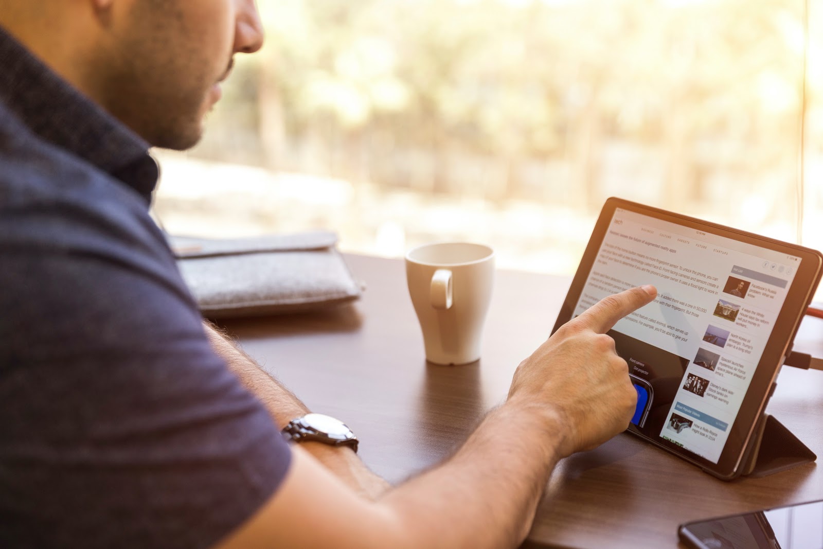 Homem mexendo em um tablet, que está em cima de uma mesa de madeira. Ao lado do tablet, há uma caneca de café, um celular e uma pasta.
