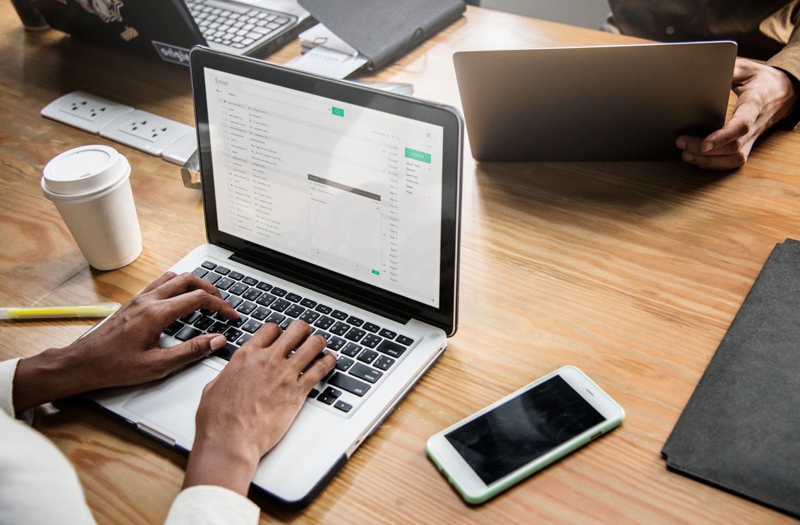 A imagem mostra as mãos de uma pessoa negra sob um laptop prata. A tela está aberta na caixa de e-mail e a pessoa parece estar digitando uma nova mensagem.