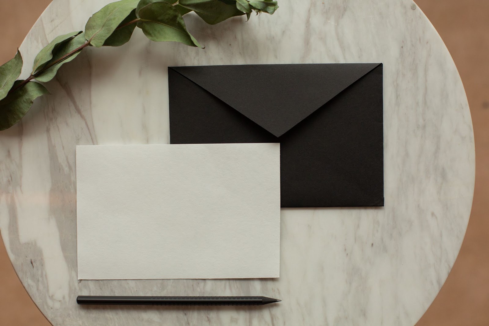 A imagem mostra uma papel branco, um envelope preto de carta, uma caneta preta e um ramo com folhas verdes sobre uma superfície de mármore circular.