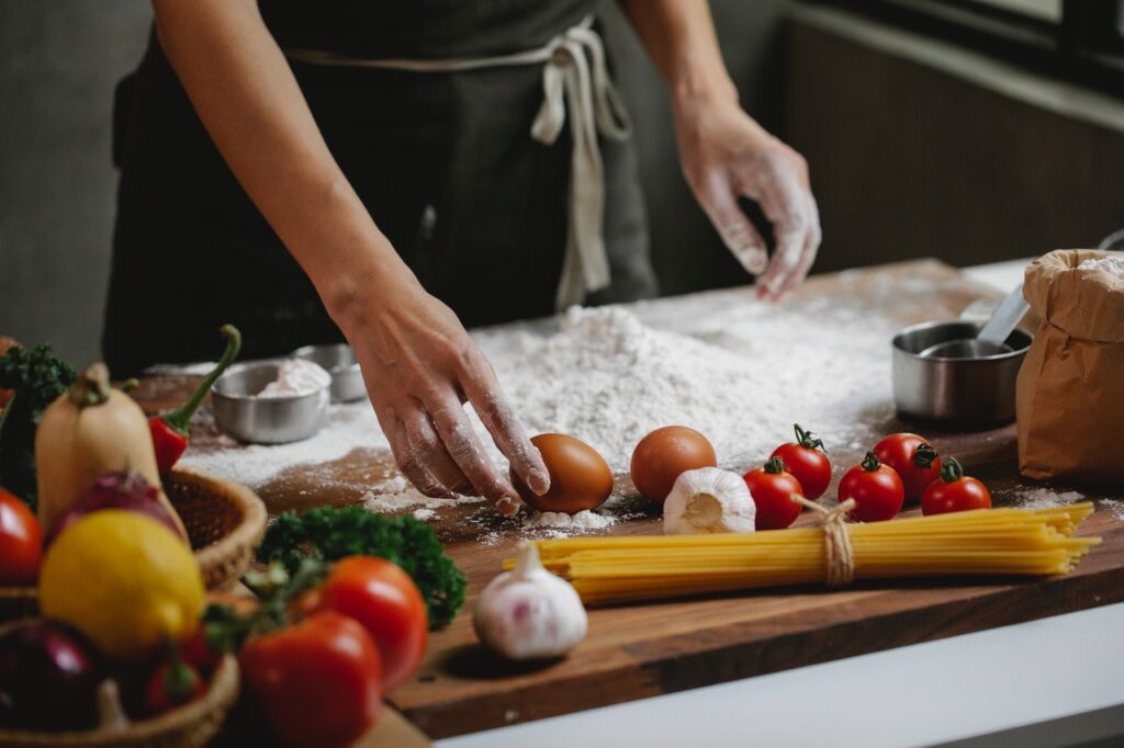 A imagem mostra, em destaque, o tronco de uma pessoa em frente a uma bancada de cozinha com alguns ingredientes. Nela, é possível ver um pouco de farinha de trigo, a mão direita da pessoa segurando um ovo marrom, ingredientes como tomate, alho, pimenta e um pouco de espaguete, ainda sem cozinhar. 