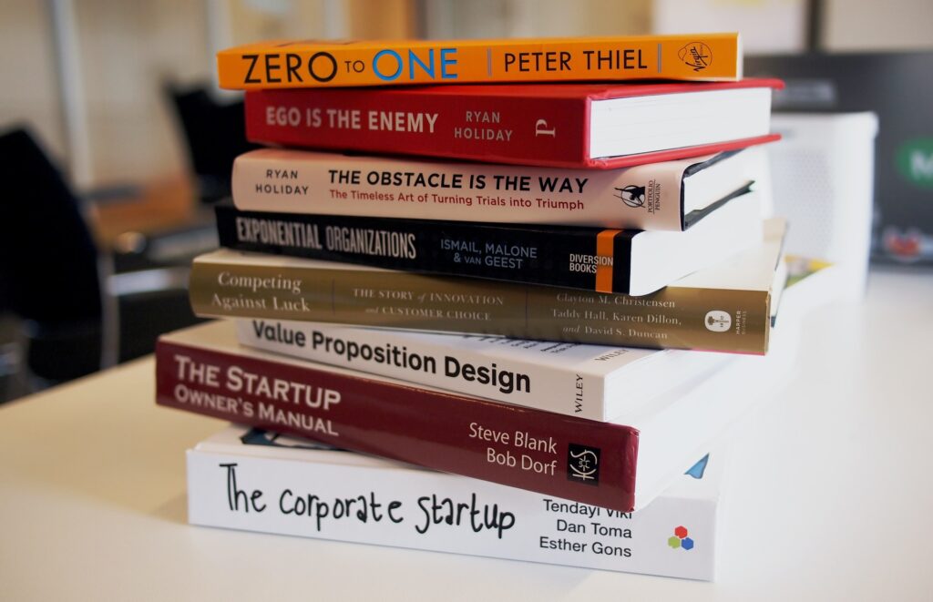 A imagem mostra oito livros empilhados sobre uma mesa branca. É possível ver a lombada deles, nas quais estão seus títulos escritos em inglês: “Zero to one l”; “Ego is a enemy”; “The obstacle is the way ”; “Exponential organizations”; “Competing against luck”; “Value Proposition Design”; “The Startup owner’s manual”; “The corporate startup”. 