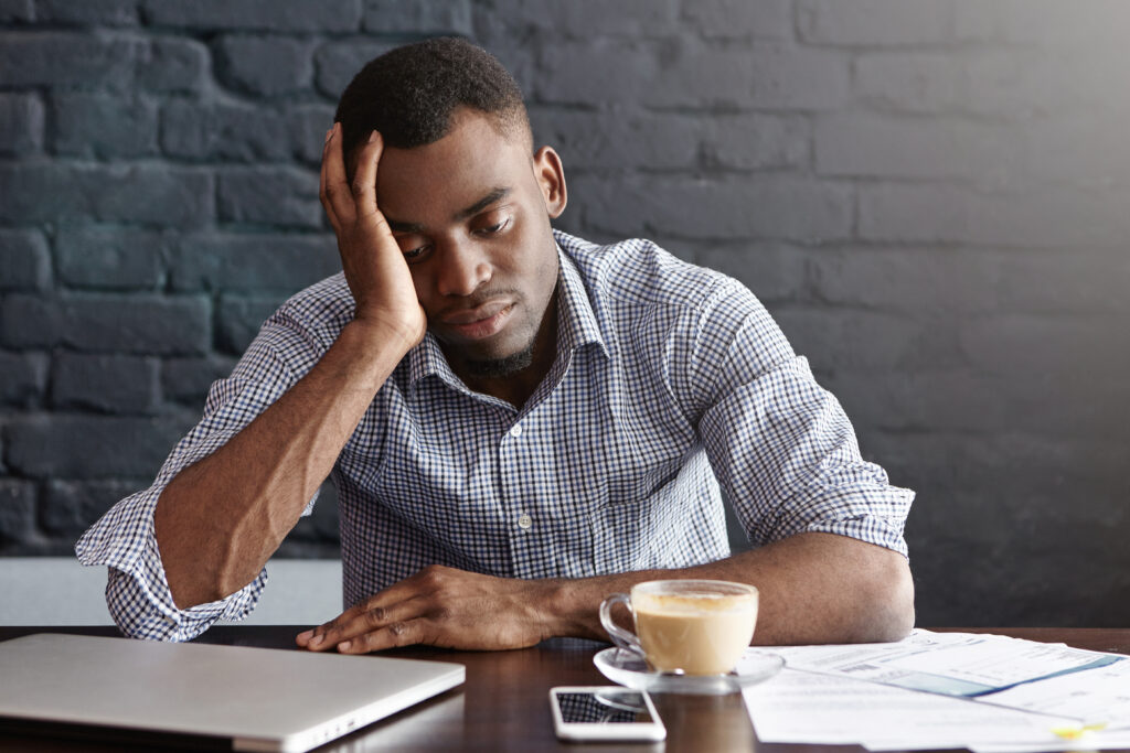 A imagem mostra um homem negro de cabelo preto sentado em frente a uma mesa de escritório. Ele veste uma camisa xadrez e está com a mão direita apoiada na cabeça, como um sinal de preocupação, e olha para baixo. Em frente a ele, na mesa, há uma xícara de café, um laptop prateado fechado, um celular branco e alguns papéis. 