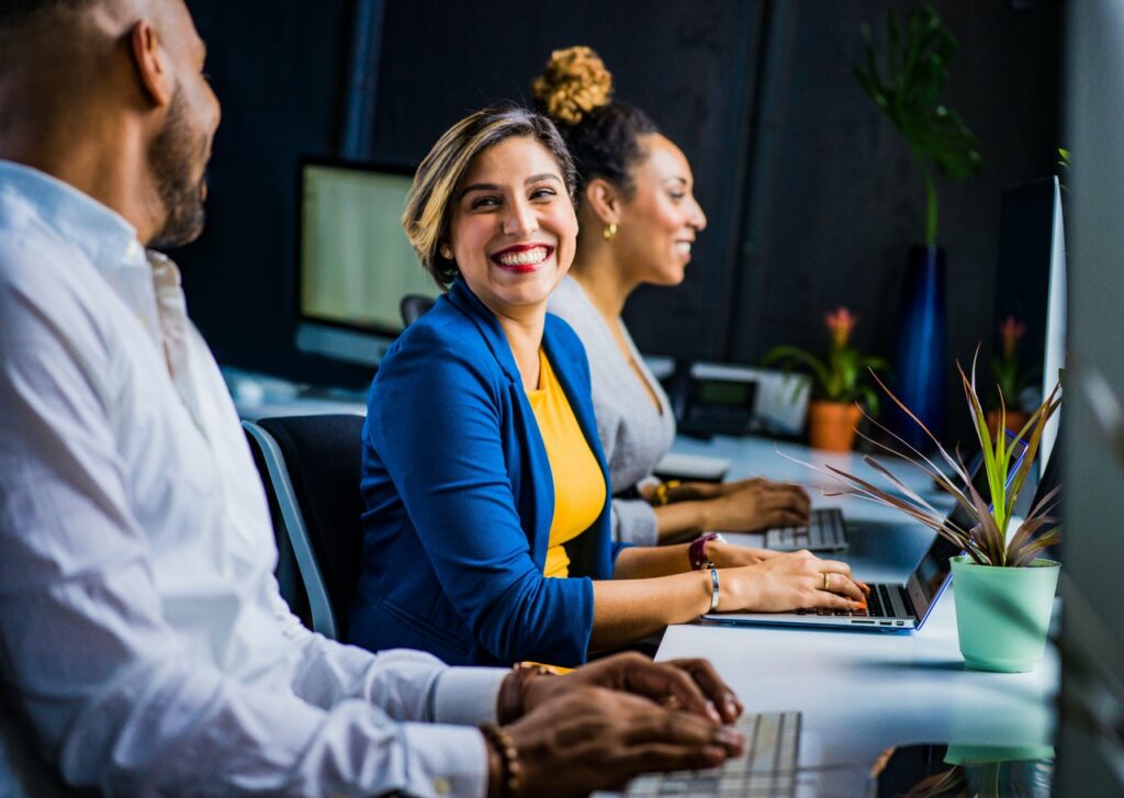 A imagem mostra um homem e duas mulheres em frente a uma mesa de escritório branca com computadores em cima dela. Em destaque na foto está uma mulher branca, de cabelo castanho com mechas loiras, cortado na altura da nuca. Ela está sorrindo e olhando para o homem ao seu lado, que está de lado para a imagem.