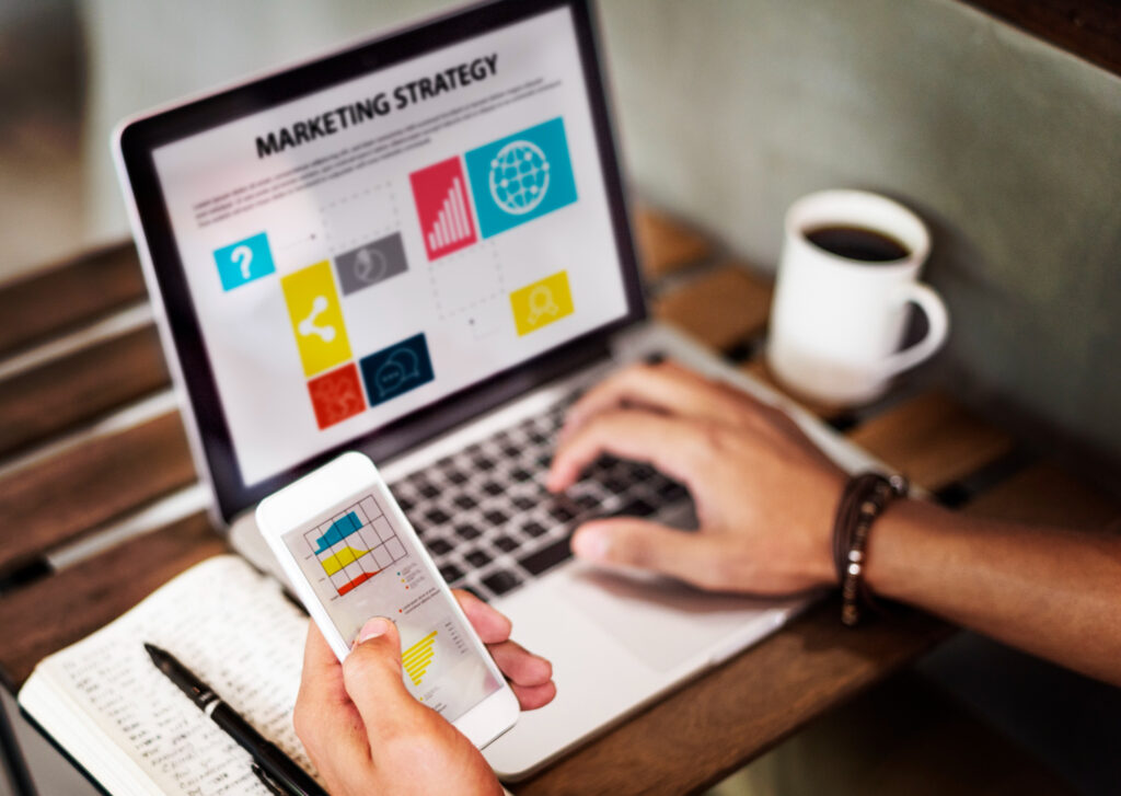 A imagem mostra, em destaque, a mão esquerda de uma pessoa segurando um celular branco, no qual é possível ver alguns gráficos coloridos. No lado direito da imagem, é possível ver um notebook prata com a frase “Marketing strategy” em destaque. A mão direita da pessoa está apoiada no teclado do notebook.