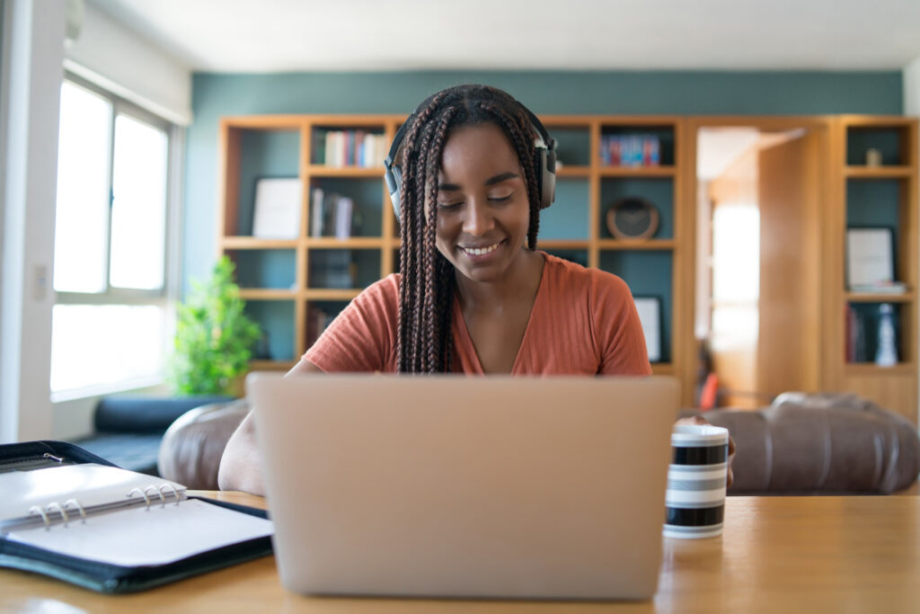 A imagem mostra uma mulher negra em frente a um laptop prata, que está em cima de uma mesa de madeira. Ela possui o cabelo trançado, usa um headset, olha para a tela do computador e sorri. Ao fundo, é possível ver uma estante de madeira que possui formato de nichos.