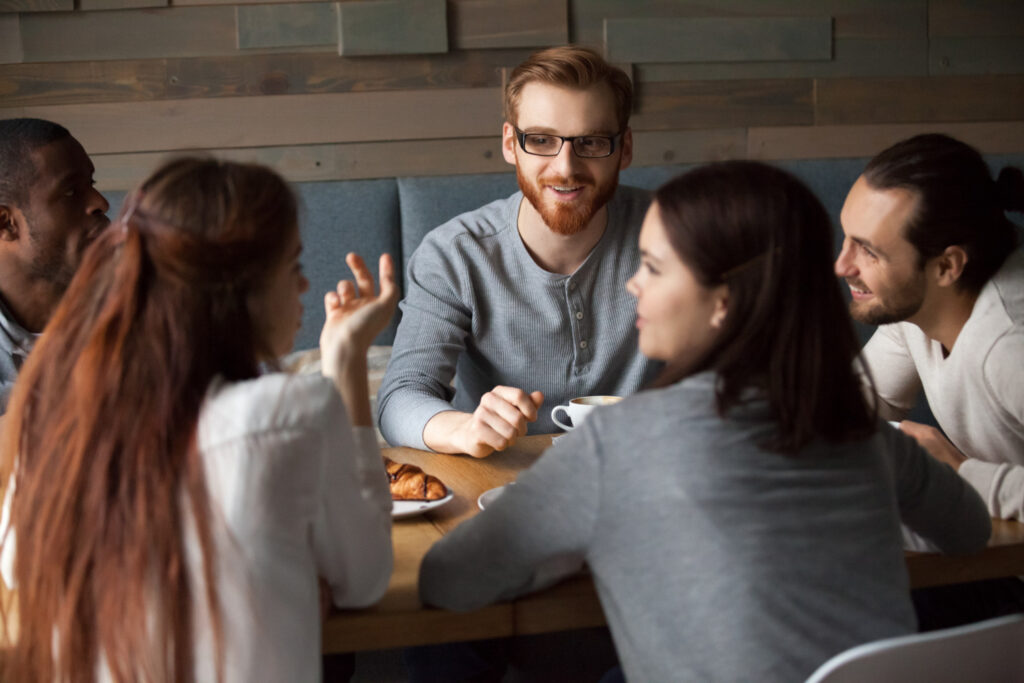 A imagem mostra um grupo de pessoas reunido em torno de uma mesa de madeira na qual é possível ver um prato com um croissant e uma xícara de café. Da esquerda para a direita, há um homem negro, ao lado dele há um homem branco ruivo, outro homem branco, de cabelo castanho-escuro preso em um coque. Ao lado desse homem, de costas para a imagem, há uma mulher branca de cabelo castanho-escuro liso na altura dos ombros e outra mulher branca ruiva. Eles parecem estar fazendo uma pausa, o que ajuda na motivação no trabalho.