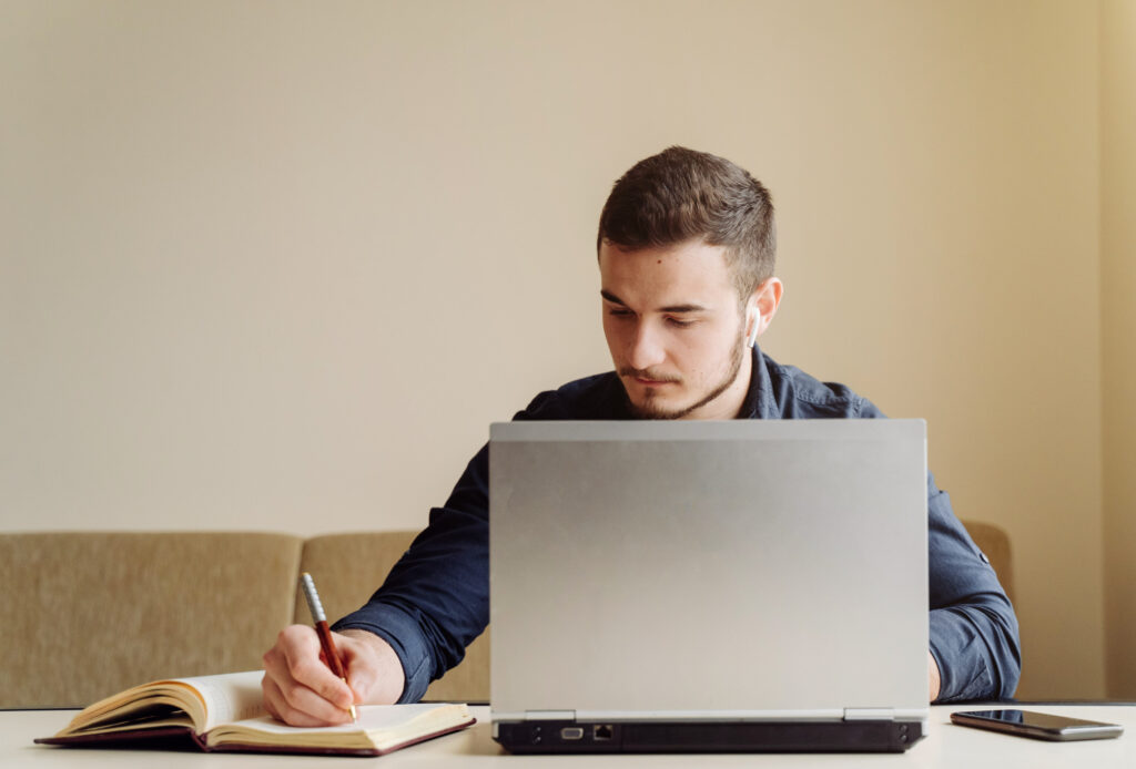 Um homem branco, de barba por fazer e cabelo curto, vestindo uma camiseta azul, está sentado em um sofá, enquanto mexe em um laptop prateado, apoiado sobre uma mesa. O homem está com uma caneta na mão direita, fazendo anotações em um caderno. Um celular também está sobre a mesa.