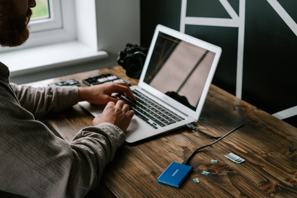 Um homem branco, com barba cheia, está sentado, mexendo em um laptop prateado, apoiado sobre uma mesa de madeira. Ao redor do laptop, sobre a mesa, estão um HD externo, alguns cartões de memória e seus adaptadores, e uma câmera fotográfica.