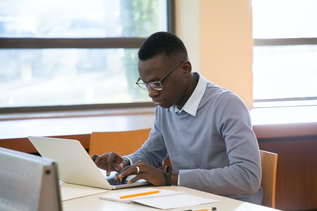 Um homem negro, de cabelo crespo escuro, usando óculos, e vestindo uma camiseta azul-clara e uma smartband, está mexendo em um laptop prateado. Ao lado do laptop, estão um caderno de anotações e um lápis. O homem está sentado em um ambiente bem iluminado pela luz natural que entra das janelas ao seu lado.