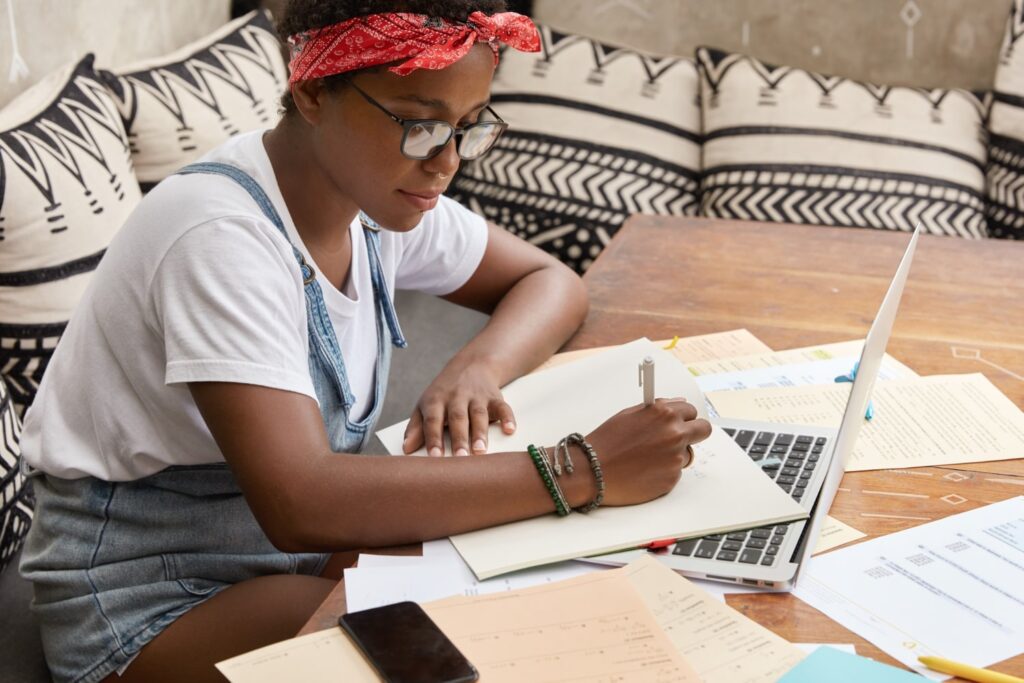 Uma mulher negra, usando óculos e com bandana na cabeça, está sentada em um sofá, fazendo anotações em um caderno. O caderno está apoiado sobre um laptop prateado que, por sua vez, está sobre uma mesa de madeira. Ainda sobre a mesa, ao redor do laptop, é possível ver um celular e vários papéis.