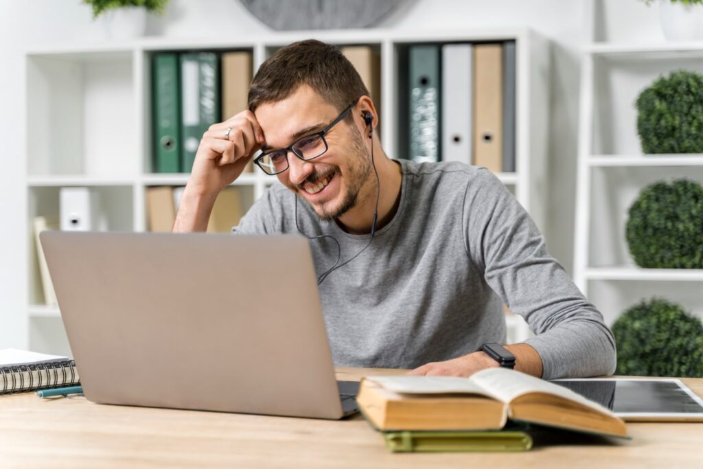 Um homem branco, de óculos e fones de ouvido, está olhando para a tela de um laptop prateado e sorrindo. O laptop está sobre uma mesa, onde também estão dois livros, um caderno de anotações, uma caneta e um tablet. Ao fundo, é possível ver uma estante.