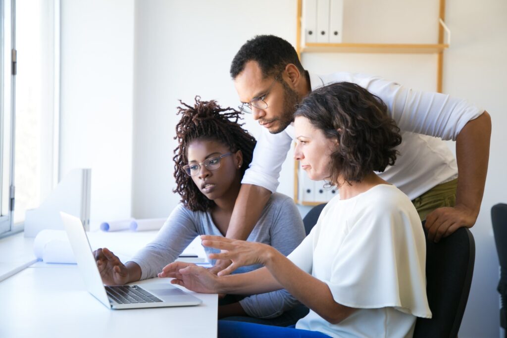 Três pessoas estão no canto direito da imagem e olham para um laptop prata que está em uma mesa branca. É possível ver uma mulher negra de cabelos trançados, um jovem negro de cabelo crespo preto e uma mulher branca de cabelo castanho ondulado.