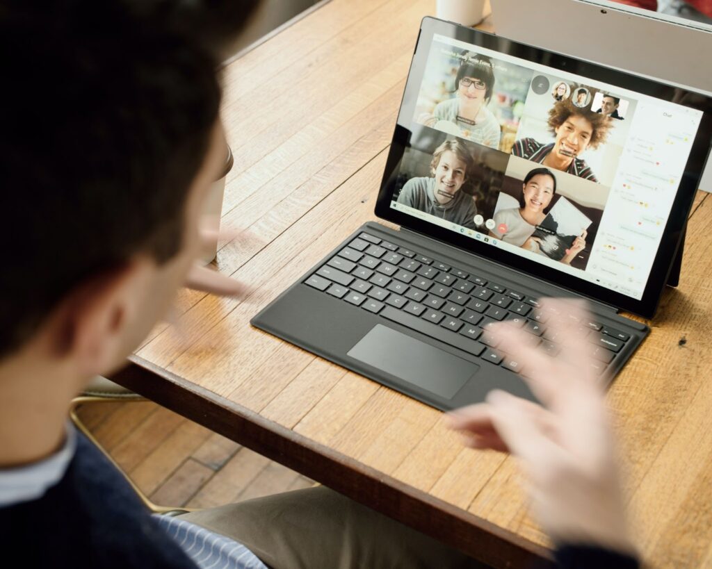 A imagem mostra um homem branco gesticulando em frente a um notebook preto slim, no qual é possível ver uma videoconferência em curso. Na chamada, é possível ver outras quatro pessoas. Da esquerda para a direita, dos quadros de cima para os de baixo, há uma mulher asiática com óculos e franja, um homem negro de cabelo crespo, um homem branco loiro e uma mulher asiática que segura um livro.
