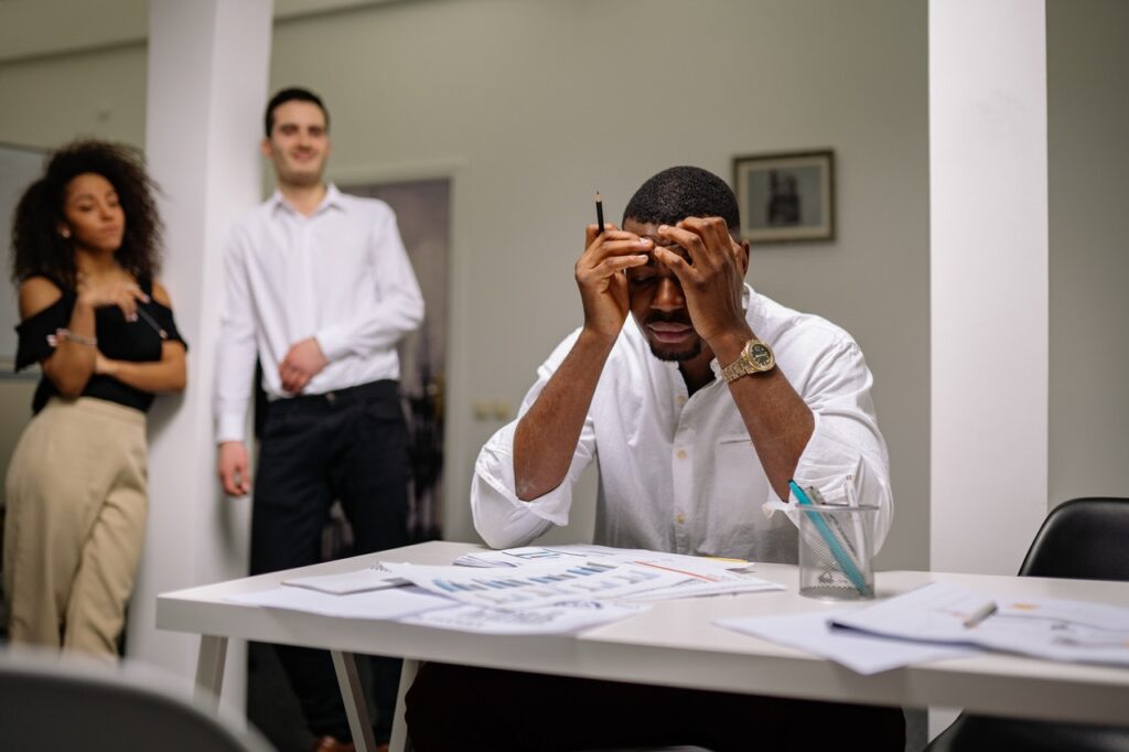 Um homem negro está sentado em frente a uma mesa de escritório, está com as mãos na cabeça e seu rosto apresenta uma expressão triste. Ao fundo há uma mulher negra e um homem branco, que olham para o homem negro. 