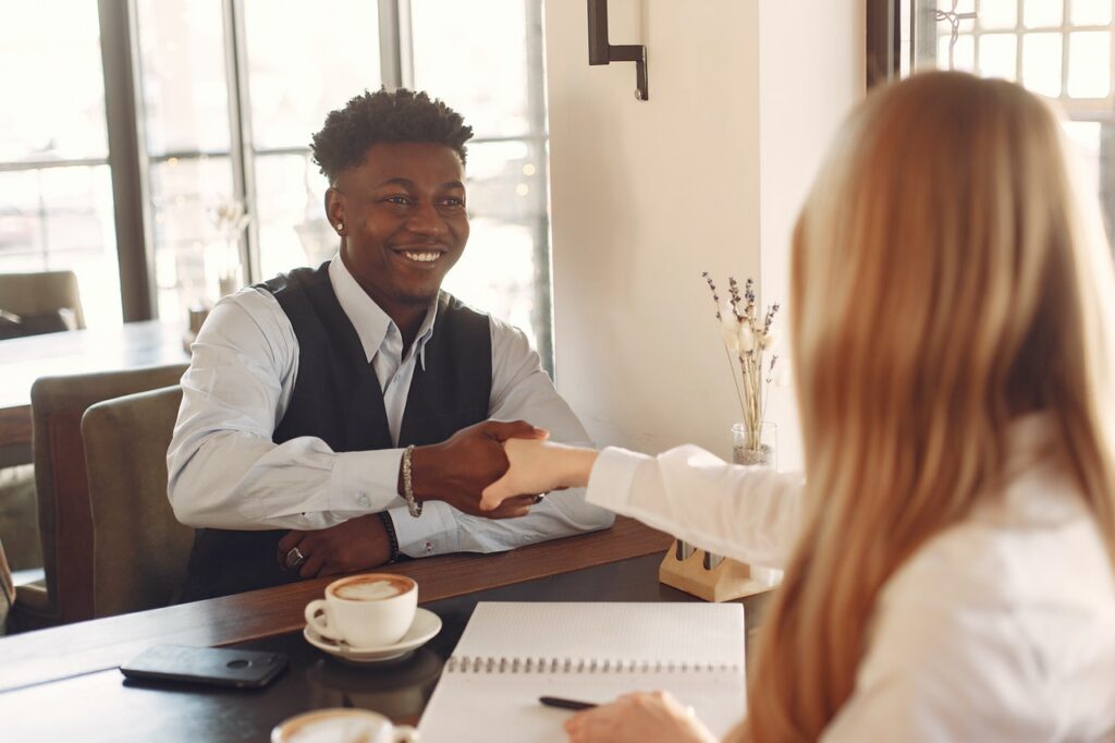 Um homem negro, de cabelo cacheado preto, está sentado em frente a uma mesa e segura a mão de uma mulher branca, ruiva, de cabelo liso, que aparece de costas para a imagem.