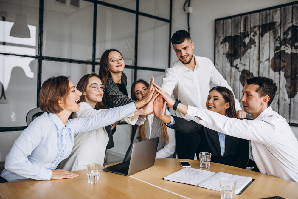 Um grupo de sete pessoas, formado por cinco mulheres brancas de cabelo liso e dois homens brancos, está reunido em volta de uma mesa de escritório. Todos aparecem com um dos braços esticados ao centro da mesa e tocam as mãos, como se estivessem comemorando uma conquista. 