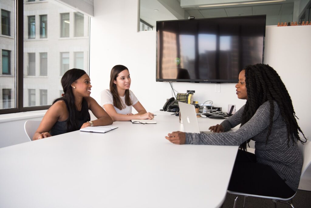 Três mulheres, duas delas negras e a outra branca, estão sentadas em uma sala, conversando. A mulher negra, à direita, está mexendo em um laptop prateado e aparenta ser uma recrutadora. A mulher branca e a outra mulher negra, ambas à esquerda, estão com cadernos de anotações, e parecem ser candidatas a uma vaga de emprego. Todas as paredes da sala são brancas e há uma grande janela atrás das candidatas. Na outra parede, é possível ver uma televisão. A mesa e as cadeiras também são brancas.