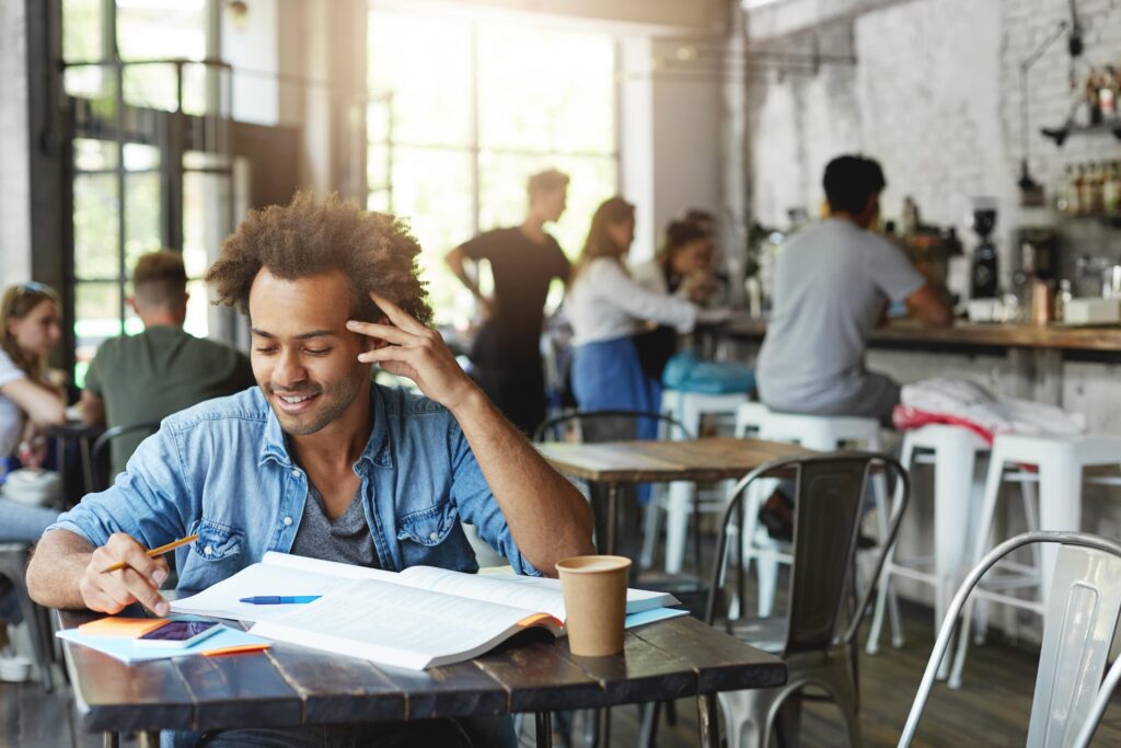 Um homem negro, de cabelo castanho-claro crespo, está sentado em uma mesa de uma cafeteria e mexe no celular. Em sua mesa estão um livro, um caderno, uma caneta, alguns blocos de anotações e um copo de café. Ao fundo há algumas pessoas.