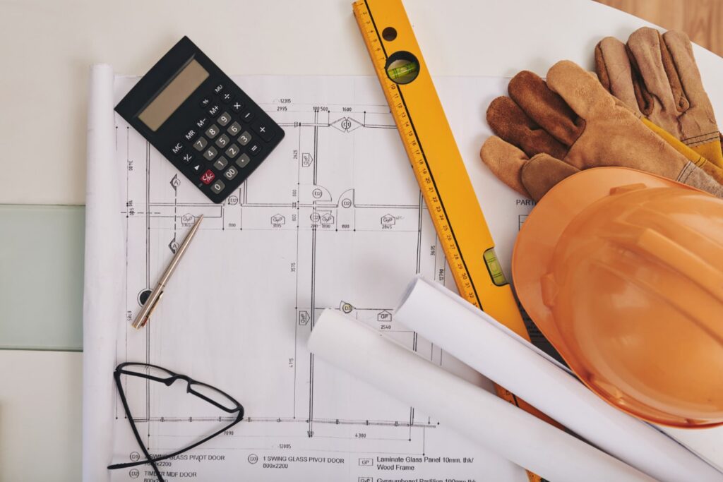 Na imagem, é possível ver vários elementos comuns na engenharia, como plantas de construções, capacete e luvas de segurança, e nível de mão. Também pode-se ver uma calculadora, uma caneta e óculos pretos.