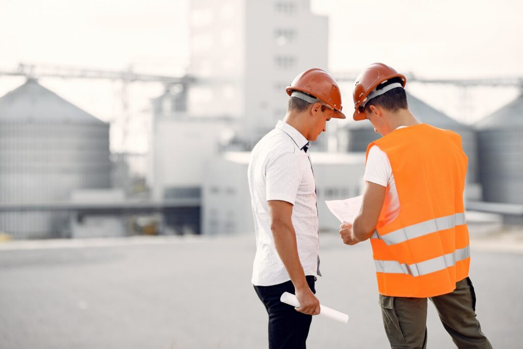 Dois homens brancos estão conversando. Os dois usam capacetes de segurança; um deles usa colete sinalizador. O de colete está mostrando um papel para o outro. Ao fundo, é possível ver uma empresa em construção.