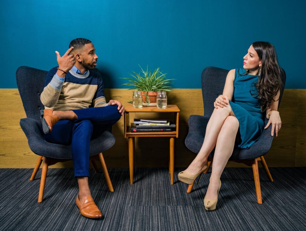 Um homem negro, de cabelo preto curto, e uma mulher branca, de cabelo castanho-escuro longo, estão conversando. Ambos estão sentados em poltronas, de pernas cruzadas. Entre as poltronas, há uma mesinha com alguns livros, uma planta e dois copos de água.