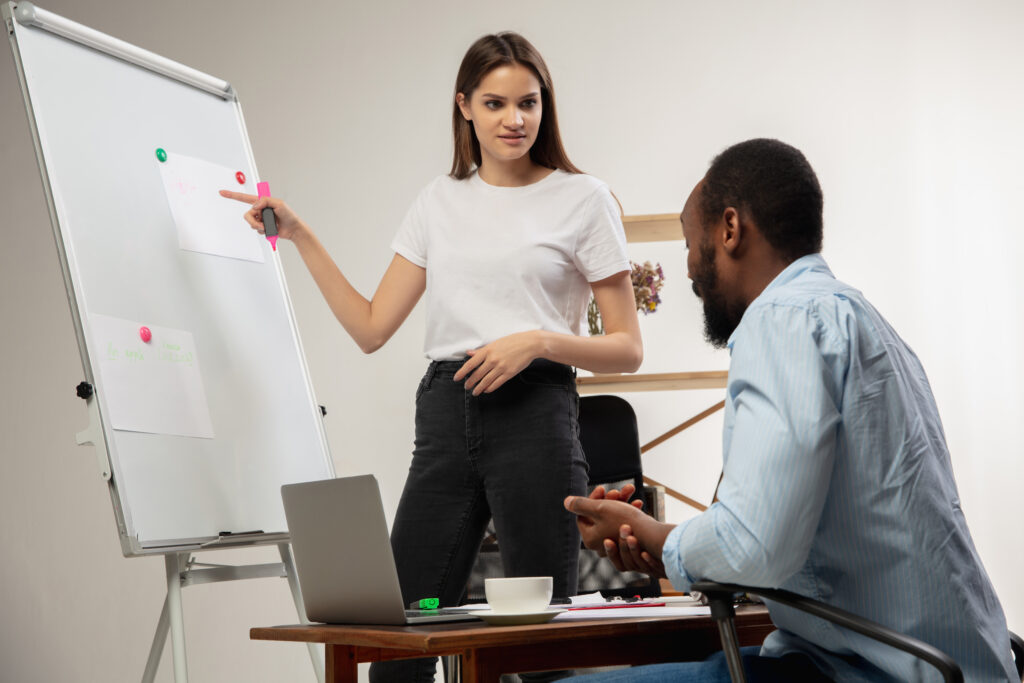 Uma mulher branca usando camiseta branca e calça preta segura uma caneta rosa e aponta para uma lousa com alguns papéis fixados. À frente da mulher, um homem negro usando camisa azul está sentado em uma cadeira em frente a uma mesa com um laptop, uma xícara e alguns papéis.