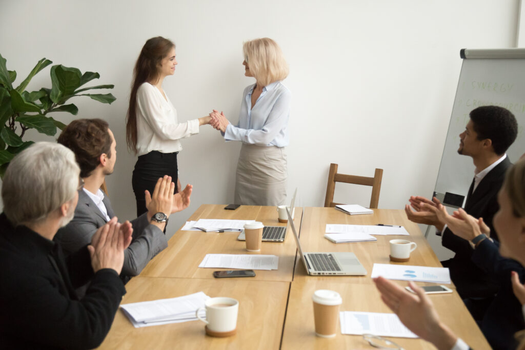 Três homens e uma mulher sentados ao redor de uma mesa de madeira com papéis, canecas e copos de café, celulares e laptops. Em pé, em frente à mesa, duas mulheres brancas, uma loira e outra morena, segurando a mão uma da outra.