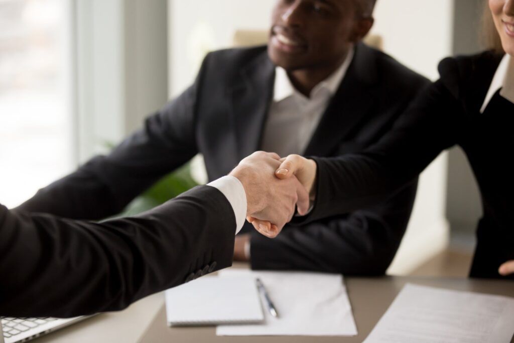 É possível ver duas pessoas brancas, sendo uma delas uma mulher, apertando as mãos. Ao fundo, um homem negro presencia a cena. Todos estão sentados ao redor de uma mesa, vestidos com roupas sociais. Há alguns papéis, uma caneta e um laptop sobre a mesa.
