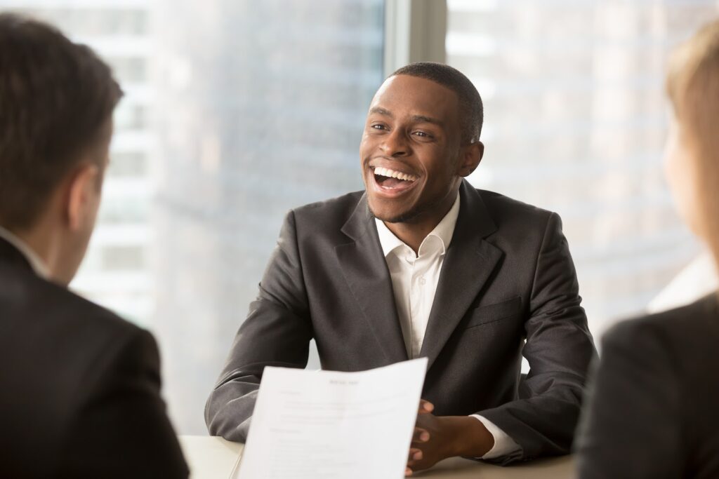 Um homem negro sorrindo e usando terno, ao centro da imagem, de frente com duas pessoas brancas desfocadas que aparecem de costas nos cantos da imagem.