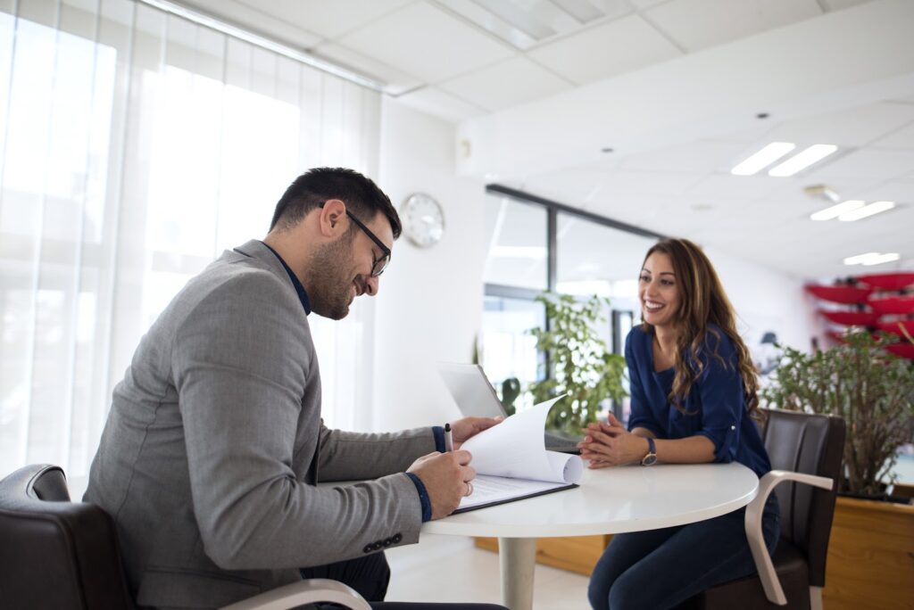 Um homem e uma mulher, ambos brancos, aparecem sentados frente a frente em uma mesa branca, no que parece ser uma entrevista de emprego. O homem está fazendo anotações em um papel; ele veste um terno cinza. A mulher usa camisa azul e calças jeans; ela está sorrindo.