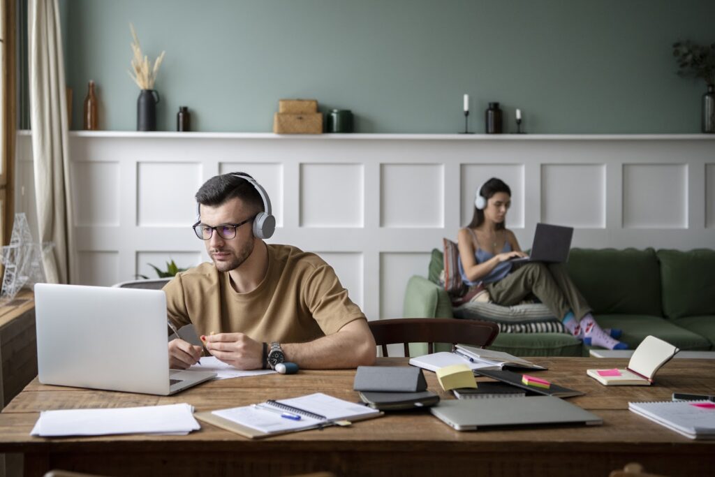 Um homem, ao lado esquerdo, mais à frente, sentado em frente a uma mesa e uma mulher, do lado direito, mais ao fundo, sentada no sofá usando laptops e headphones em uma sala. Ter consistência nos estudos é uma dica de como melhorar o listening.