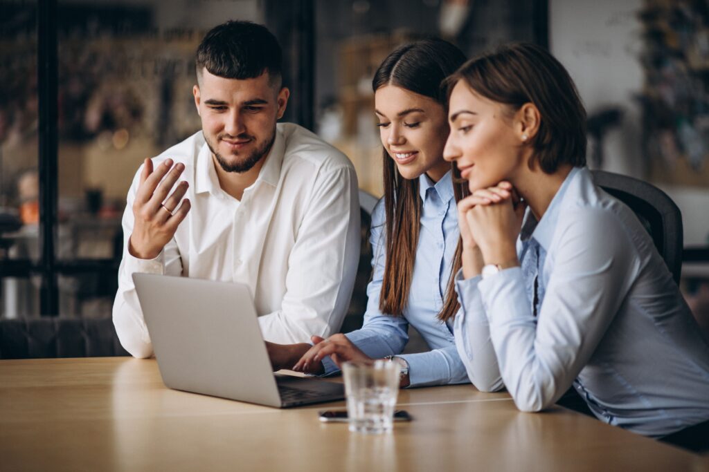 Duas mulheres e um homem usando roupas sociais e sentados em frente a um laptop prata, que está em cima de uma mesa de madeira.
